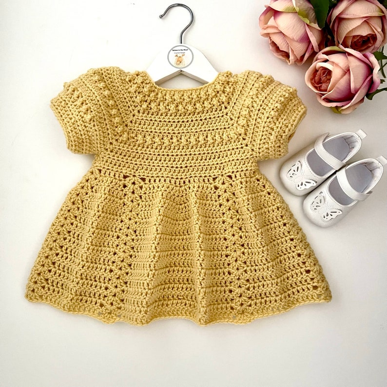 May include: A yellow crocheted baby dress with short sleeves and a scalloped hem. The dress is hanging on a white hanger with a small deer logo.