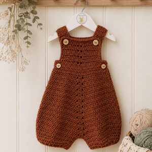 May include: A rust-colored crochet romper featuring a vertical openwork stitch pattern down the center, four light-colored wooden buttons at the shoulders and sides, and a wide leg opening, displayed on a white hanger against a paneled wall.