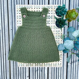 May include: A green crocheted dress with two buttons on the straps. The dress is lying on a woven mat with green plants in the background.