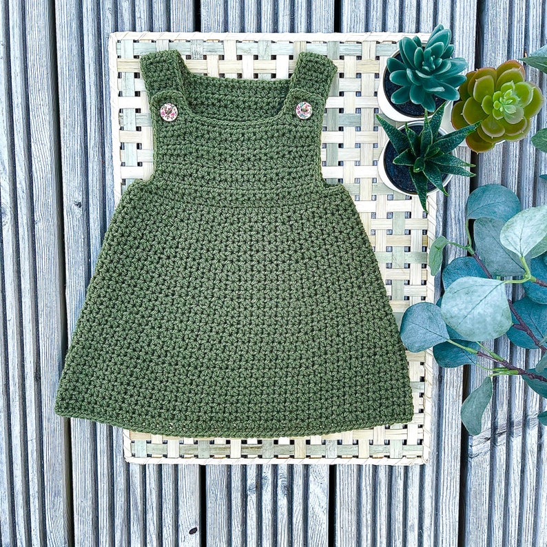 May include: A green crocheted dress with two buttons on the straps. The dress is lying on a woven mat with green plants in the background.