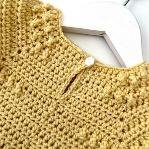 May include: A yellow crocheted garment with a white button closure. The garment has a ruffled neckline and a textured stitch pattern.