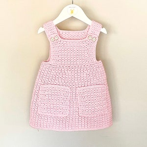 May include: A pink crocheted baby dress with two pockets and button accents.