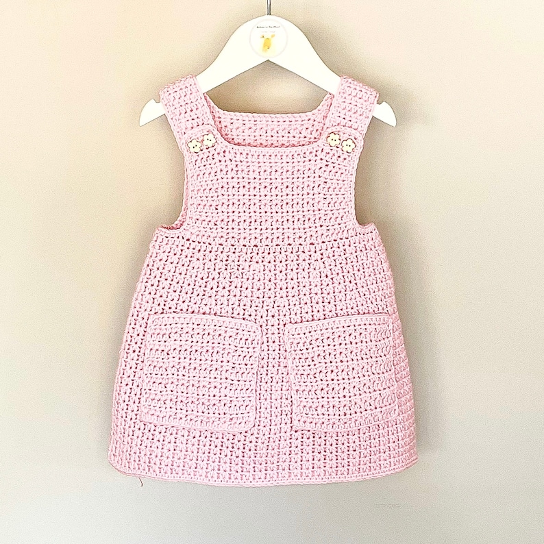 May include: A pink crocheted baby dress with two pockets and button accents.