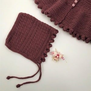 Könnte beinhalten: Ein Set aus gehäkelter Babyhaube und -schal in einem tiefen Burgunderrot. Die Haube hat einen Bindfaden und der Schal einen gewellten Rand.