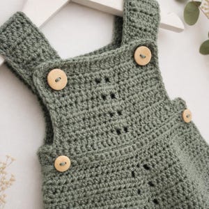 May include: A sage green crocheted romper featuring a textured stitch pattern, wide shoulder straps, and four light-colored wooden buttons attached at the front and sides. The garment is displayed on a white hanger, showcasing the yarn construction.