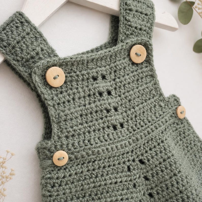 May include: A sage green crocheted romper featuring a textured stitch pattern, wide shoulder straps, and four light-colored wooden buttons attached at the front and sides. The garment is displayed on a white hanger, showcasing the yarn construction.