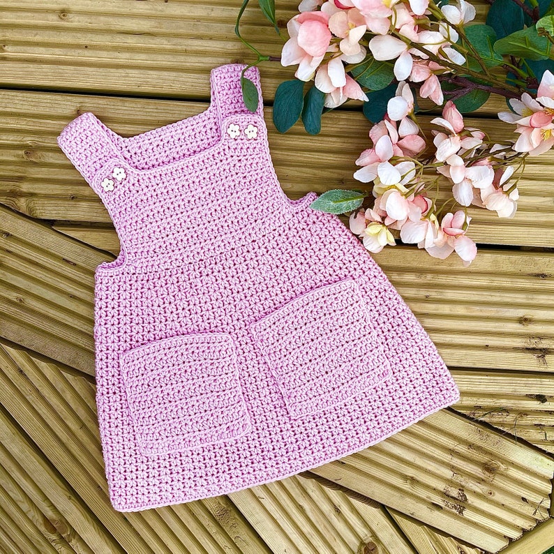 May include: A pink crocheted dress with two pockets and buttons on the straps.