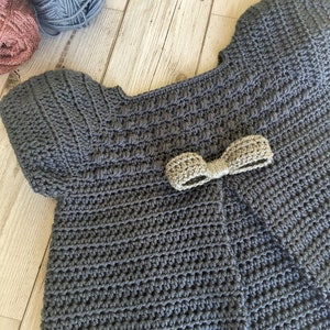 May include: A gray crocheted baby sweater with a gold bow on the front.