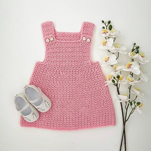 May include: A pink crocheted baby dress with white buttons. The dress has a sleeveless design and a gathered skirt.