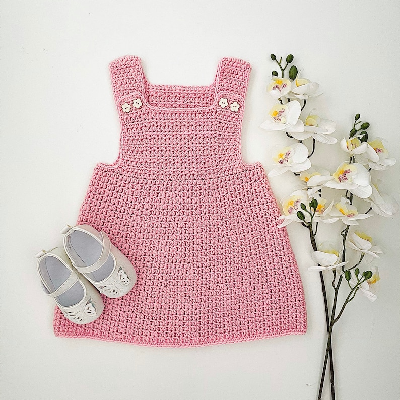 May include: A pink crocheted baby dress with white buttons. The dress has a sleeveless design and a gathered skirt.