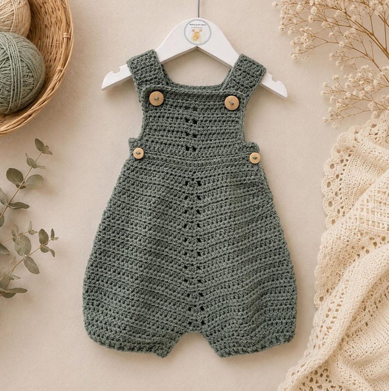May include: A sage green crocheted romper featuring a sleeveless design with wide shoulder straps. The garment includes four small, round, light-colored wooden buttons at the front, two on the straps and two on the sides, with a vertical textured stitch pattern running down the center.