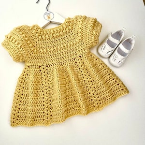 May include: A yellow crocheted baby dress with short sleeves and a scalloped hem. The dress is hanging on a white hanger with a small deer logo.