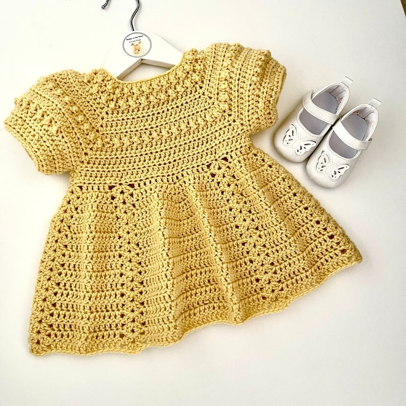 May include: A yellow crocheted baby dress with short sleeves and a scalloped hem. The dress is hanging on a white hanger with a small deer logo.