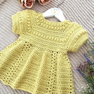 May include: A yellow crocheted baby dress with short sleeves and a ruffled hem. The dress is hanging on a white hanger.