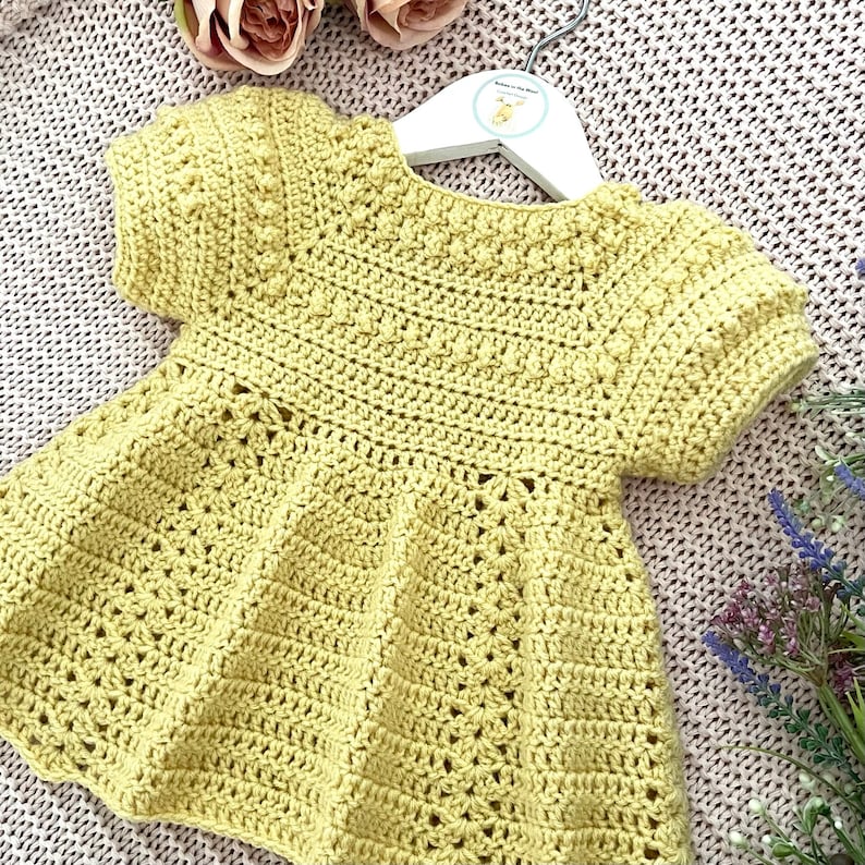 May include: A yellow crocheted baby dress with short sleeves and a ruffled hem. The dress is hanging on a white hanger.