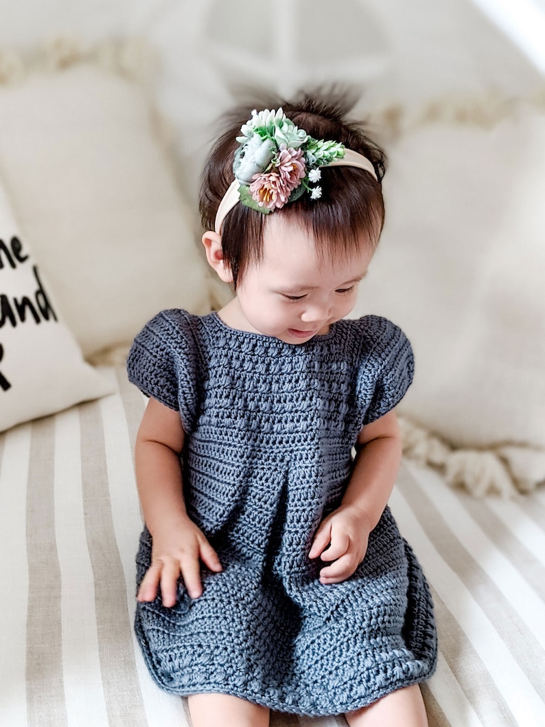 May include: A young child wearing a blue crocheted dress and a headband with a floral design. The child is sitting on a striped bed.