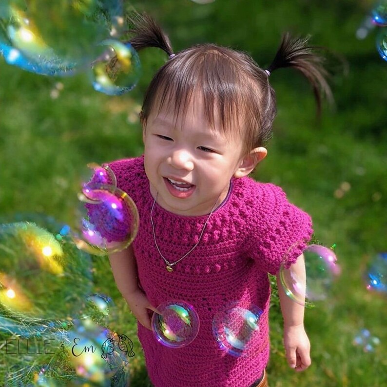 May include: A young girl wearing a pink crocheted dress with a silver necklace smiles while playing with bubbles outdoors.