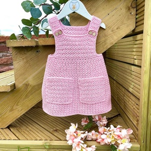 May include: A pink crocheted dress with two pockets and wooden buttons. The dress is hanging on a wooden hanger.