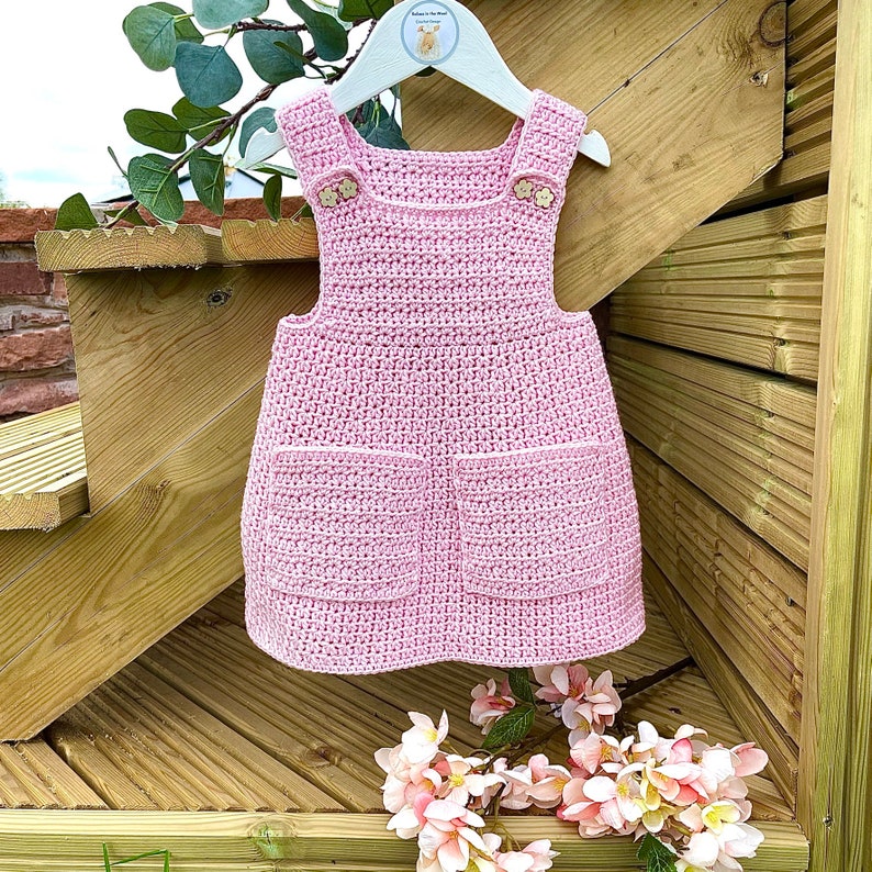 May include: A pink crocheted dress with two pockets and wooden buttons. The dress is hanging on a wooden hanger.