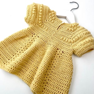 May include: A yellow crocheted baby dress with short sleeves and a round neckline. The dress has a delicate pattern and a slightly flared skirt.