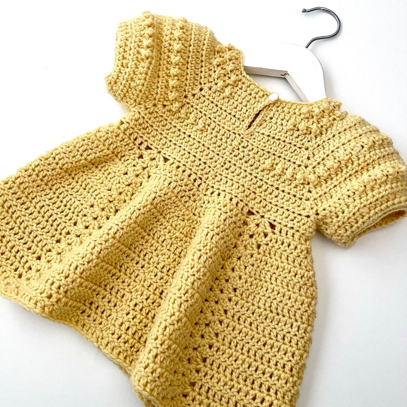 May include: A yellow crocheted baby dress with short sleeves and a round neckline. The dress has a delicate pattern and a slightly flared skirt.