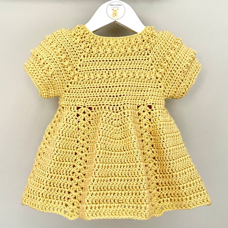 May include: A yellow crocheted baby dress with short sleeves and a flared skirt. The dress has a delicate crocheted pattern throughout.