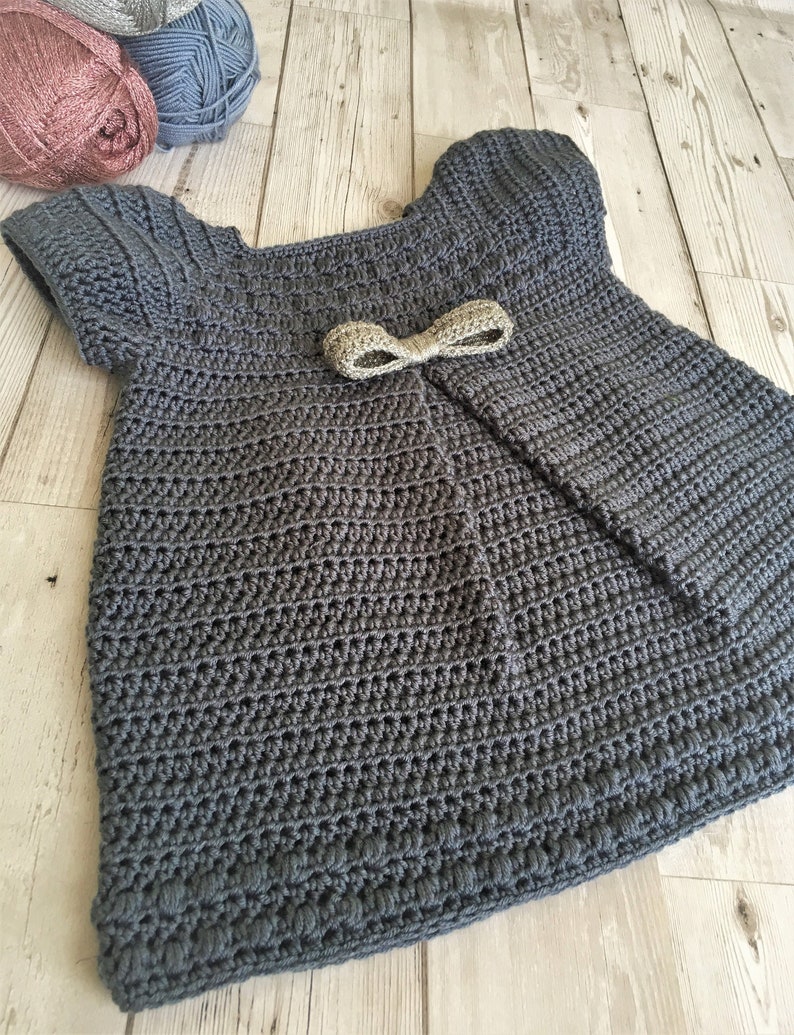 May include: A gray crocheted baby dress with short sleeves and a gold bow. The dress has a gathered skirt and a scalloped hem.