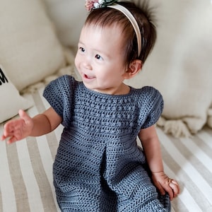 May include: A baby girl wearing a blue crocheted dress with a floral headband. The dress has a simple design and is perfect for a special occasion.