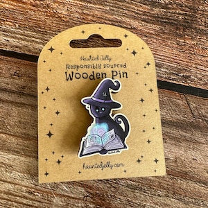 May include: Wooden pin of a black cat in a purple witch hat, reading a spell book. The pin is on a brown card with the text "Haunted Jelly Responsibly sourced Wooden Pin" and "hauntedjelly.com". The pin has a spooky, Halloween theme.