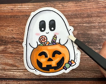 Może przedstawiać: Naklejka o tematyce Halloween z białym duchem z czarnymi oczami i różowymi policzkami, trzymającym pomarańczową dynię wypełnioną słodyczami. Dynia ma wyrzeźbioną twarz. Naklejka jest trzymana przez pęsetę. Widoczny jest tekst "Haunted Jelly".