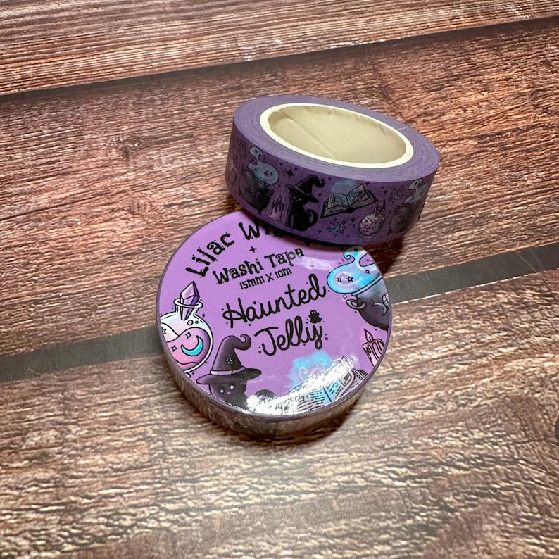 May include: Two rolls of washi tape with a Halloween theme. The tape is purple with illustrations of witch hats, potion bottles, and other spooky elements. The text "Lilac Washi Tape" and "Haunted Jelly" are visible.