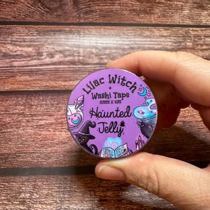 May include: A roll of Lilac Witch washi tape, 15mm x 10m, featuring a purple background with witch-themed illustrations. The design includes a witch's hat, potion bottles, a cauldron, and the words "Haunted Jelly".