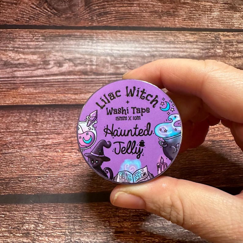 May include: A roll of Lilac Witch washi tape, 15mm x 10m, featuring a purple background with witch-themed illustrations. The design includes a witch's hat, potion bottles, a cauldron, and the words "Haunted Jelly".