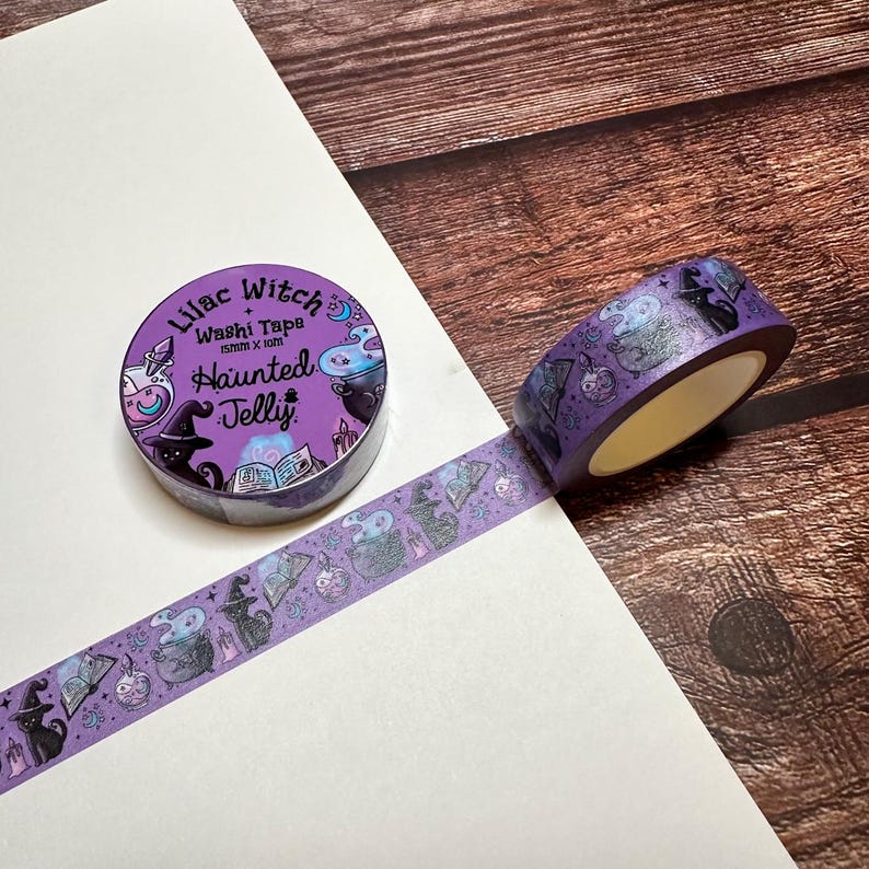May include: A roll of purple washi tape with a Halloween theme. The tape features illustrations of witch hats, cauldrons, cats, and the text "Lilac Witch Haunted Jelly". The tape is 15mm x 10m.