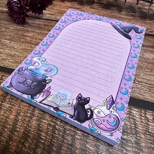 May include: A lined lavender notepad with a witchy theme. The notepad features illustrations of a cauldron, a black cat, a potion bottle, and a witch's hat. The border is decorated with crescent moons and stars.
