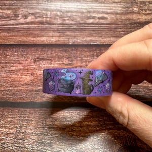May include: Purple washi tape featuring Halloween-themed illustrations. The tape has images of a cauldron, a black cat wearing a witch hat, a spell book, and potion bottles. The tape is held by a person's hand.