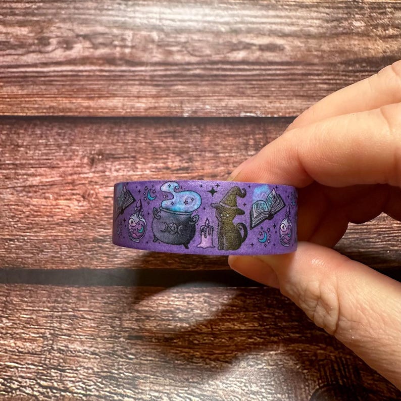 May include: Purple washi tape featuring Halloween-themed illustrations. The tape has images of a cauldron, a black cat wearing a witch hat, a spell book, and potion bottles. The tape is held by a person's hand.