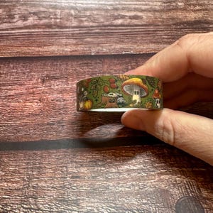 May include: A roll of decorative washi tape featuring a whimsical forest scene. The tape has a green background with illustrations of mushrooms, pumpkins, berries, and leaves. The tape is held in a person's hand.