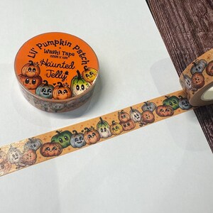 Op de afbeelding: Oranje washi tape rol met een pompoen ontwerp. De tape heeft cartoon pompoenen in verschillende kleuren en uitdrukkingen, met de tekst "Li'l Pumpkin Patch Haunted Jelly". De tape is 15 mm x 10 m.