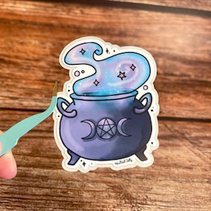 Peut inclure: Un chaudron violet avec un symbole de triple lune et de pentacle est rempli de fumée tourbillonnante bleue et violette. La fumée a des étoiles roses et un contour blanc. L'autocollant dit "Haunted Jelly".