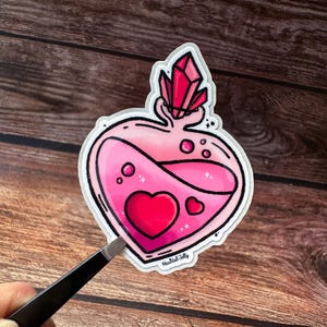 Könnte beinhalten: Ein herzförmiger Aufkleber mit einem rosa Trank-Design. Der Aufkleber zeigt ein großes rotes Herz, kleinere Herzen und einen Kristall oben. Der Trank befindet sich in einer herzförmigen Flasche mit schwarzem Umriss. Der Aufkleber hat den Text "Haunted Jelly".