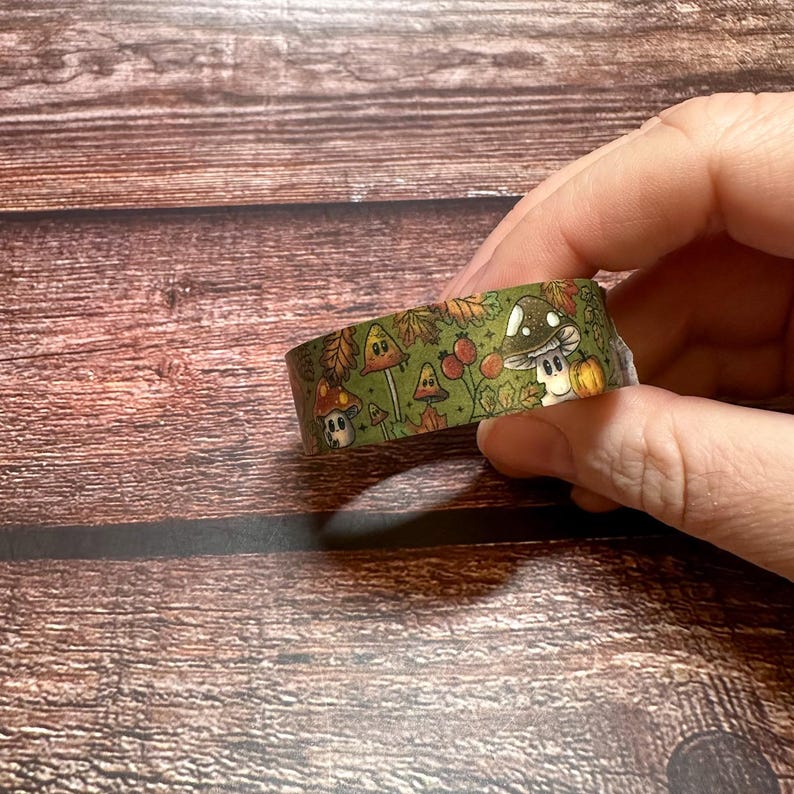 May include: Decorative washi tape featuring a whimsical mushroom design in shades of green, brown, and orange. The tape roll displays various cartoon mushrooms, leaves, and berries. The tape is held in a hand against a wooden background.