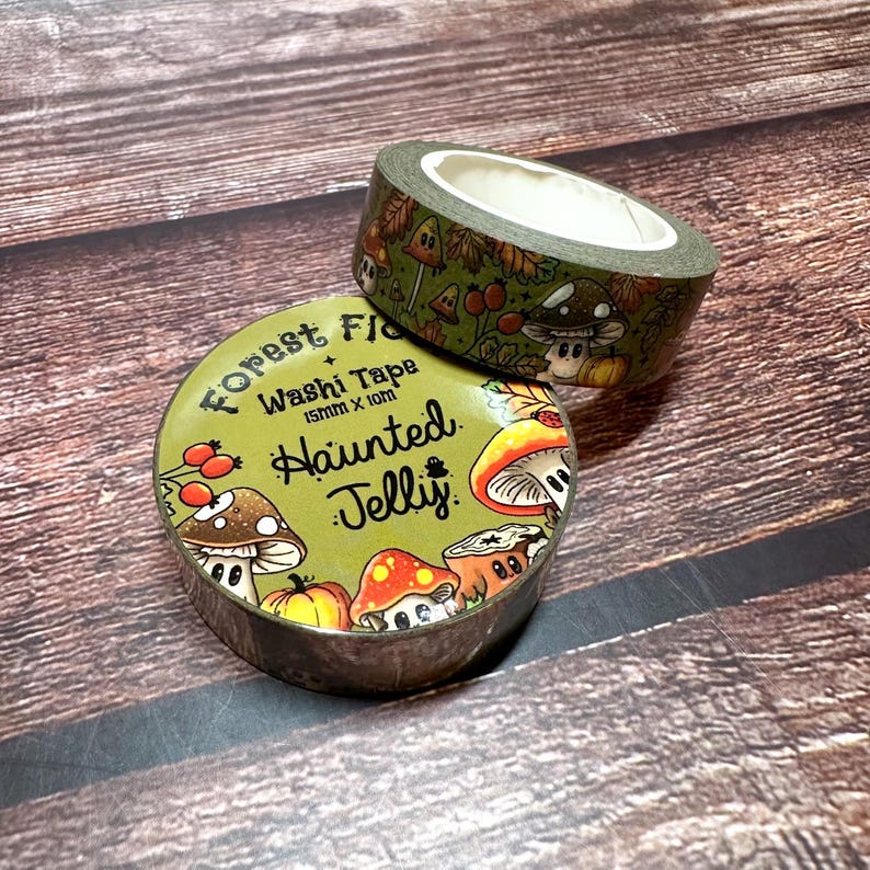 May include: Two rolls of washi tape with a forest theme. The tape features illustrations of mushrooms, berries, and leaves in shades of green, orange, and brown. One roll is stacked on top of the other. The text "Forest Flora" and "Haunted Jelly" are printed on the tape.