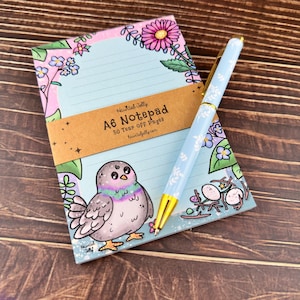 May include: A6 notepad featuring a cartoon pigeon illustration, floral border, and lined pages. A light blue pen with a floral design lies next to the notepad. The notepad has the text "Haunted Jolly A6 Notepad 50 Tear Off Pages hauntedjolly.com".