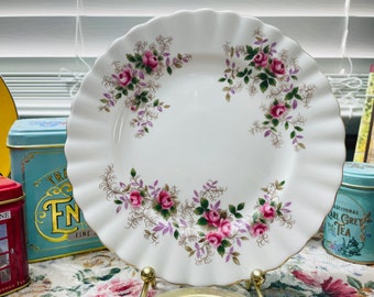May include: A white ceramic plate with a scalloped edge features a delicate floral pattern of pink roses and purple leaves. The plate is perfect for serving tea or dessert.
