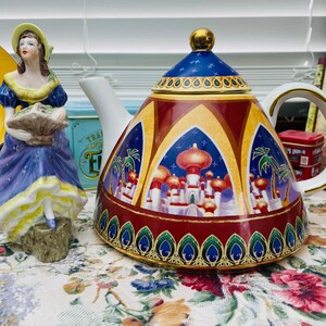 May include: A red and gold teapot with a blue and gold lid and a scene of a city with minarets and palm trees. The teapot has a gold handle and spout.