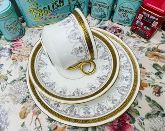 May include: A white teacup and saucer set with a gold and gray floral pattern. The set features a delicate design with a gold rim and intricate details.