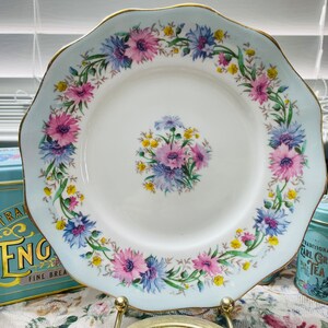 Könnte beinhalten: Ein weißer Porzellanteller mit einem blauen und weißen Blumendesign. Der Teller hat einen gewellten Rand und einen goldenen Rand. Die Blumen sind rosa, blau und gelb.