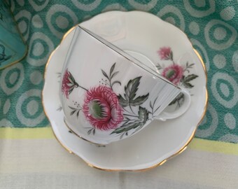 Puede incluir: Una taza de té y un platillo blancos con un diseño floral. La taza y el platillo tienen un borde dorado. El diseño floral presenta flores rosas y verdes.