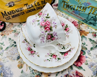 May include: A white teacup and saucer set with a floral pattern featuring pink roses and green leaves. The set has a gold trim around the edges.